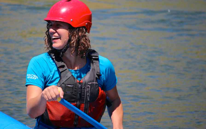 a person wearing a life jacket and helmet smiles while sitting in a raft and using a paddle